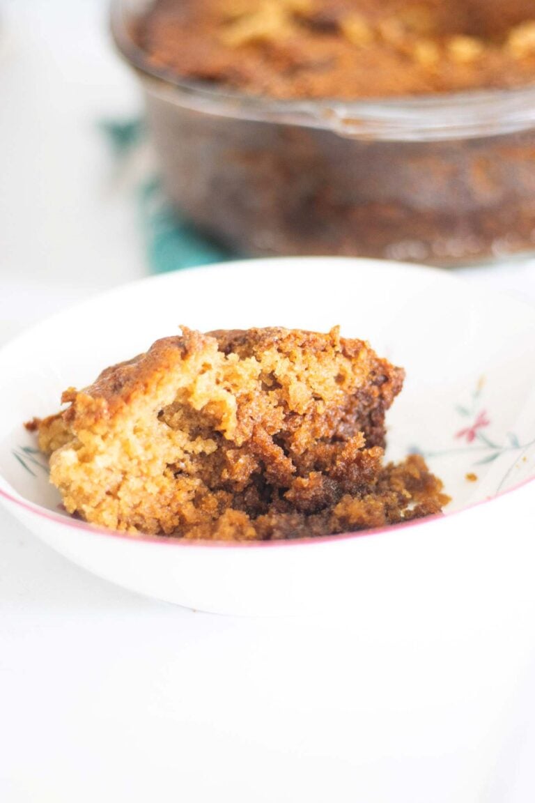 There is a slice of sourdough coffee cake in a white and floral bowl with the glass baking dish of coffee cake in the background.