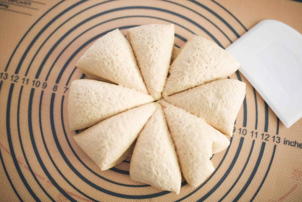 Hoagie sandwich roll dough is divided into 8 equal pieces with a white bench scraper beside them.