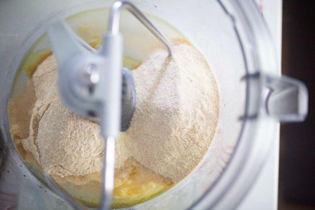 Flour is sitting on top of some liquid ingredients in a clear stand mixer bowl.