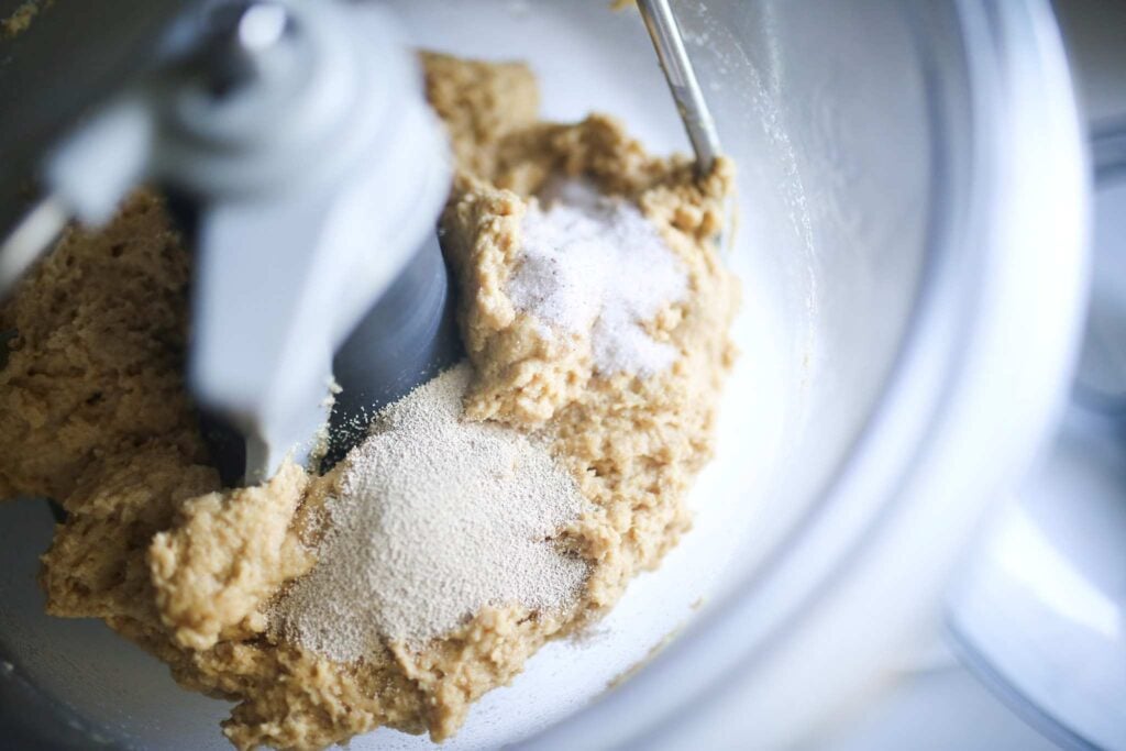 There is a pile of yeast and a pile of salt sitting atop some dough in a clear stand mixer bowl.