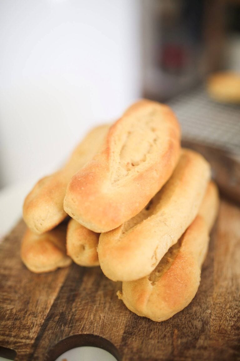 There is a stack of golden brown hoagie rolls on a dark wood cutting board.