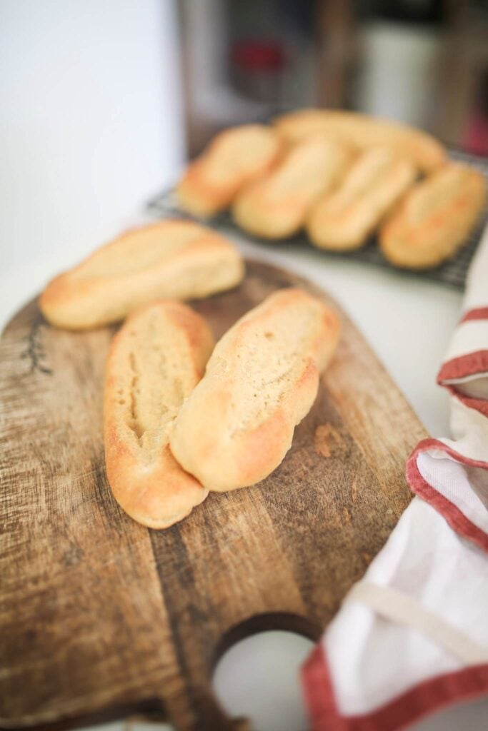 There are 2 stacked and one not stacked sandwich rolls on a cutting board with more in the background on a cooling rack.