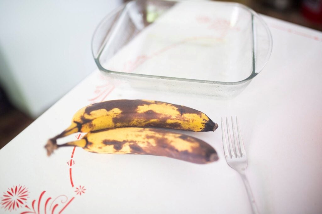 2 over ripe bananas sit on a white and red table in front of a glass share pan and beside them is a fork.