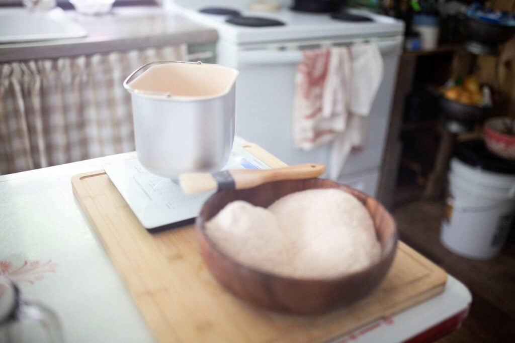 In the foreground is a wooden bowl of flour and in the background is a bread machine pan on a kitchen scale.