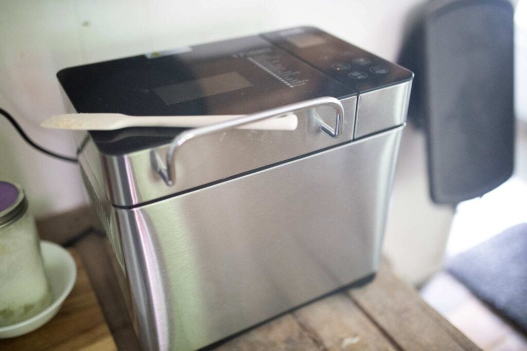 A picture of a silver bread machine with a white spatula on top.