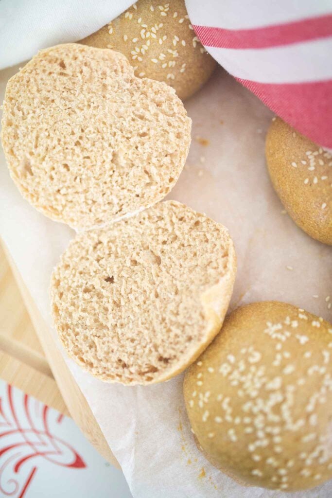 A hamburger bun cut open to show the light and airy crumb sits on a piece of parchment paper with more hamburger buns.