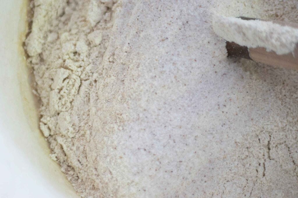 There is flour and salt on top of wet ingredients in a cream colored bowl with a dough whisk in the bowl.