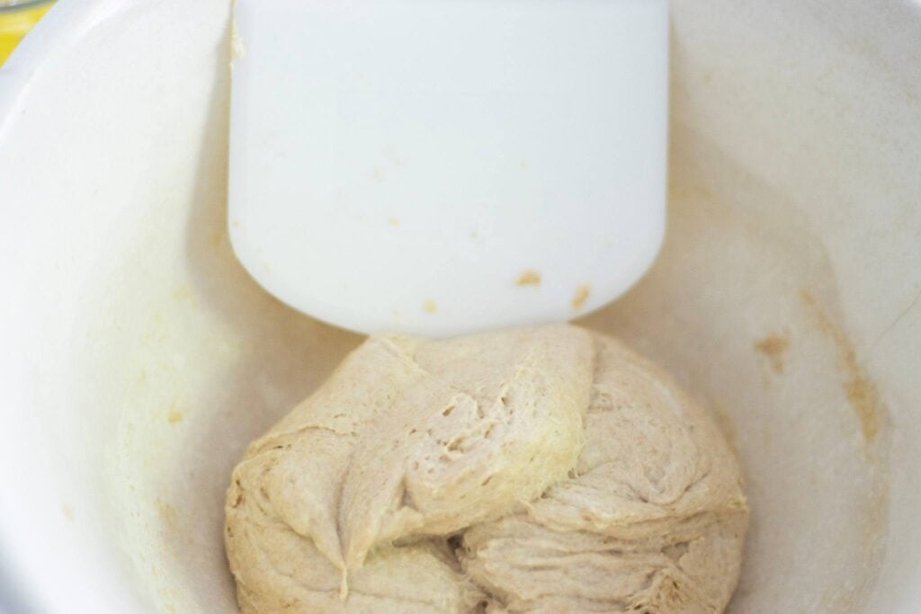 There is dough that has been folded over itself in a cream bowl with a white bowl scraper in the bowl as well.