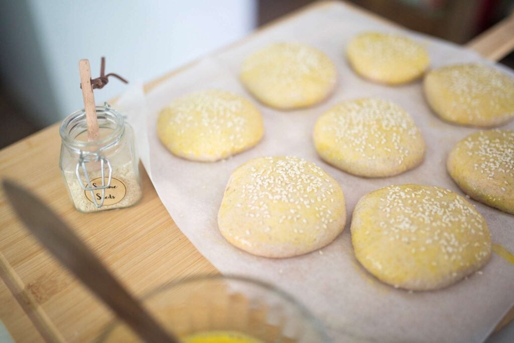 Shaped hamburger bun dough is topped with an egg wash and sprinkled with sesame seeds.