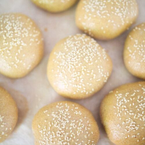 There are several hamburger buns topped with sesame seeds on a piece of parchment paper.