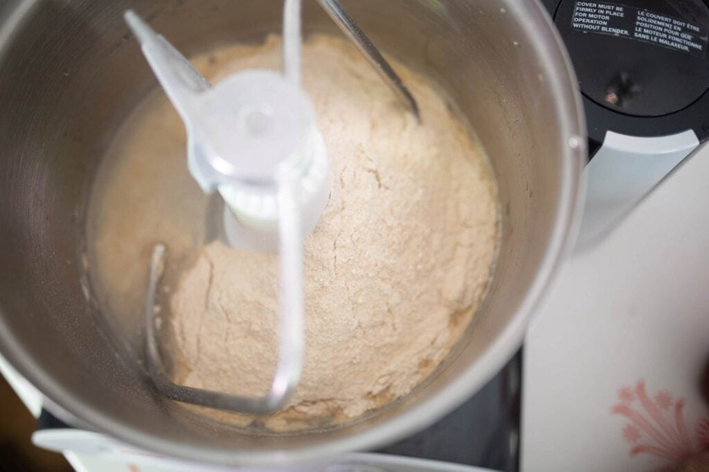 Wet and dry ingredients in a stainless steel mixer bowl with a dough hook attachment.