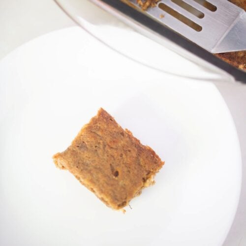 An overhead photo of a banana bar on a white plate with the pan of bars beside it with a metal spatula in it.
