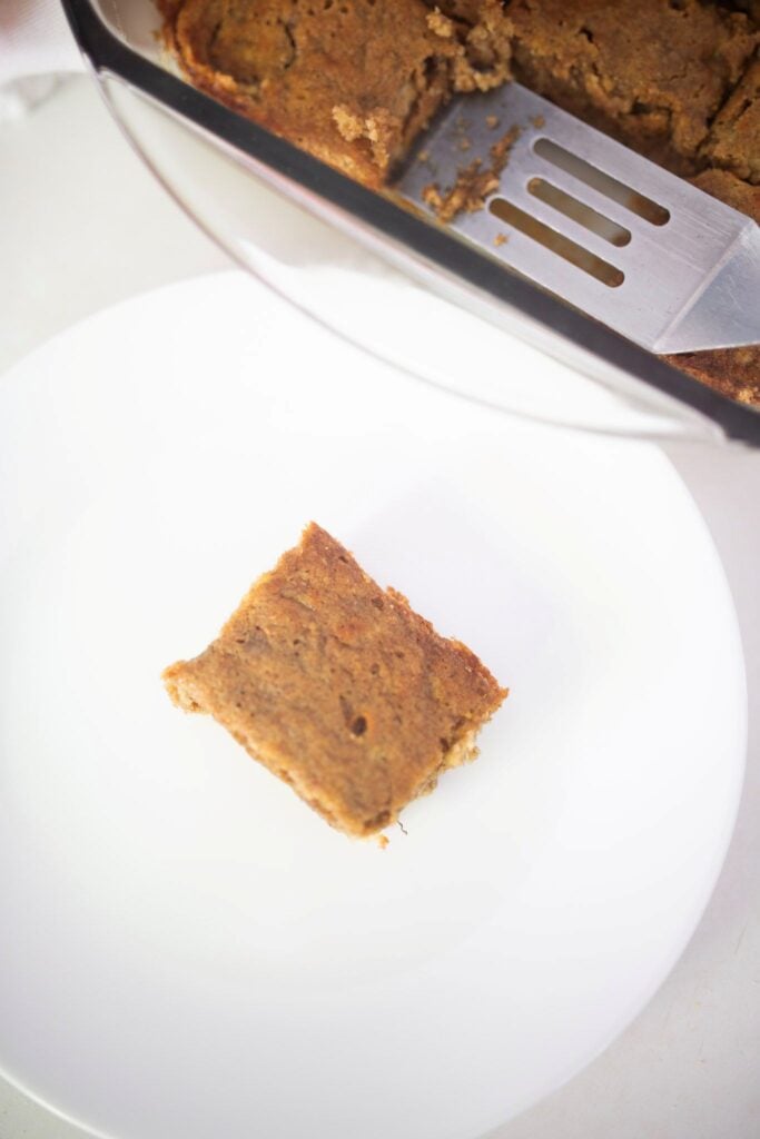 An overhead photo of a banana bar on a white plate with the pan of bars beside it with a metal spatula in it.