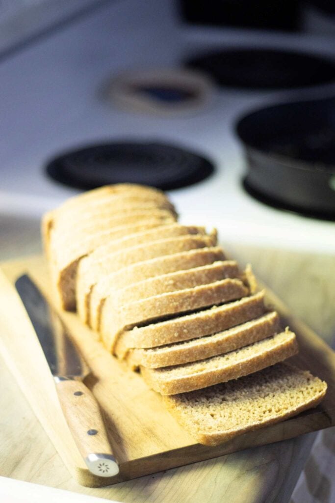 There is a sandwich loaf, sliced, on a cutting board with a bread knife. It sits on a wood counter with a white oven in the background.