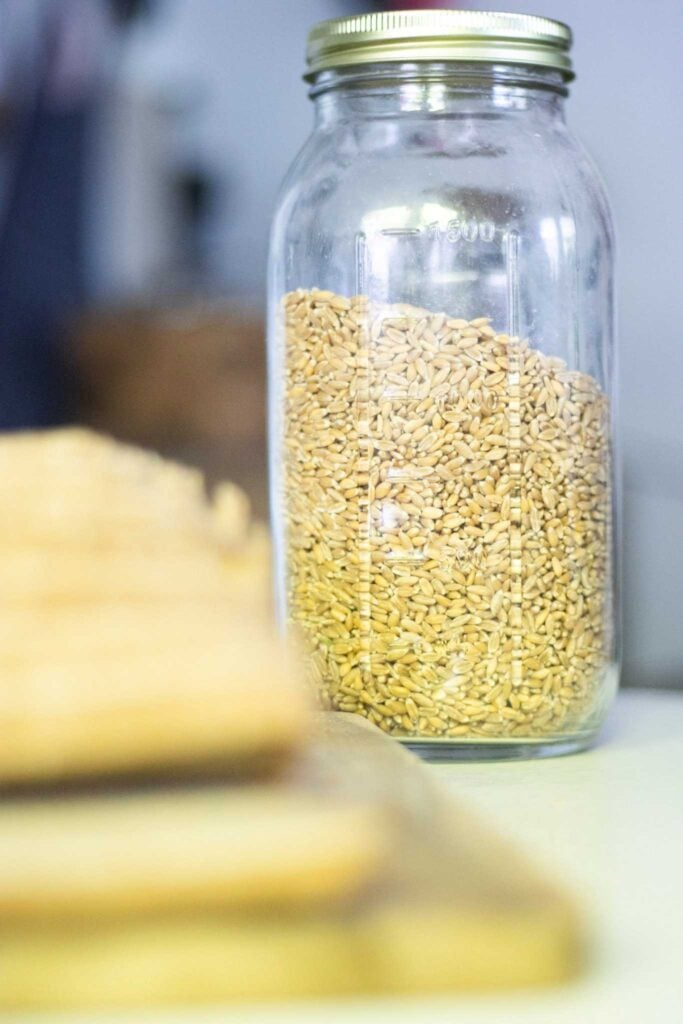 A large glass jar of wheat berries sits behind a sliced loaf of bread.