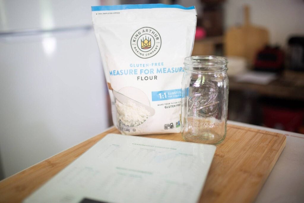 A bag of flour, mason jar, and kitchen scale sit on a cutting board.