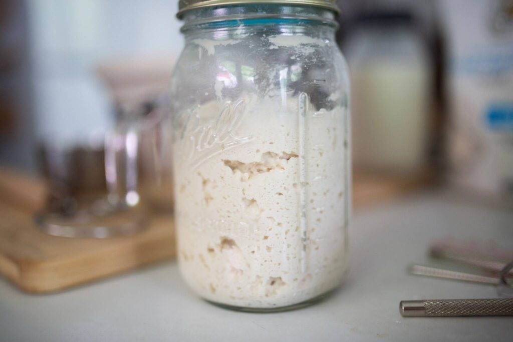 A jar sits on a counter with sourdough starter in it. It is in front of a butcher block.