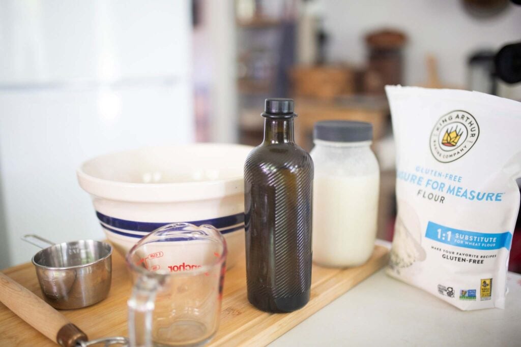 Ingredients for gluten free biscuits on a wooden cutting board. Flour, olive oil, milk, measuring cup, and mixing bowl on a cutting board.
