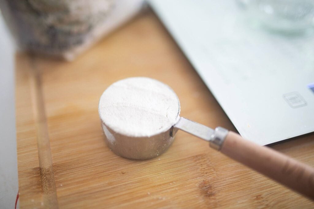 A small measuring cup full of flour sits on a wood cutting board beside a kitchen scale.
