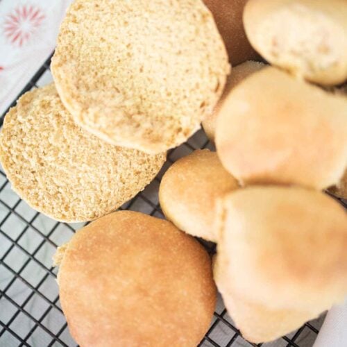 Hamburger buns and rolls on cooling rack with a bread knife and one of the buns cut open to show the crumb.