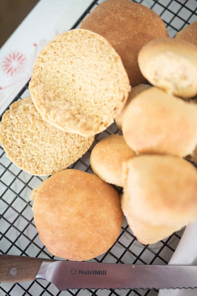 Hamburger buns and rolls on cooling rack with a bread knife and one of the buns cut open to show the crumb.