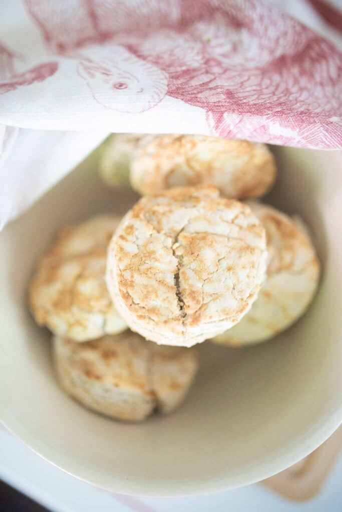 An overhead picture of a golden brown biscuit sitting on top of a pile of biscuits in a cream colored bowl with a tea towel in the background.
