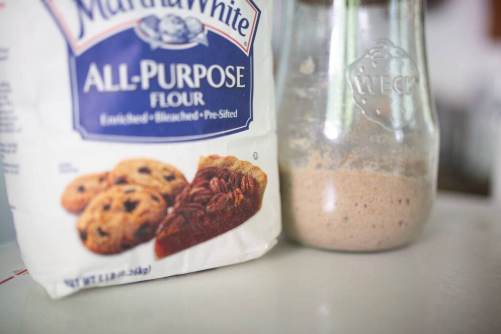 A bag of all purpose flour and a jar of sourdough starter sitting on a white table top.