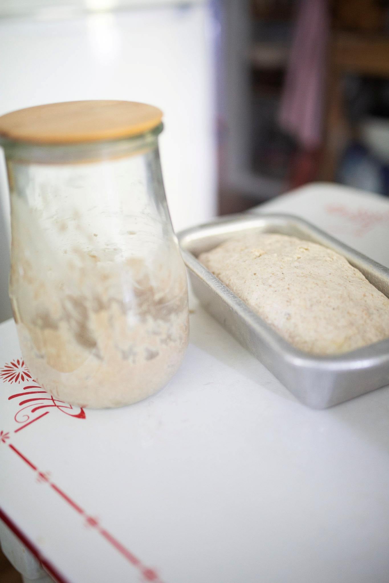 Sourdough starter in a jar with a wooden lid sits beside a loaf pan of bread dough that has risen.