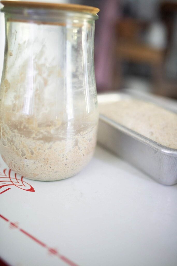 There is a jar of sourdough starter beside a loaf pan of dough.