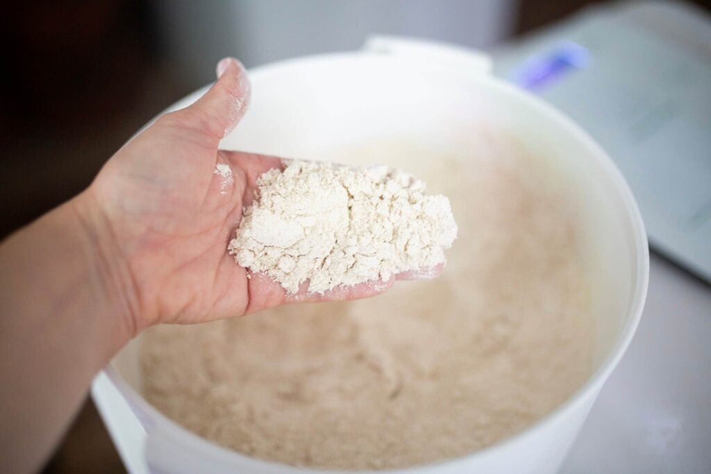 A hand holding flour over a big white bin of flour.