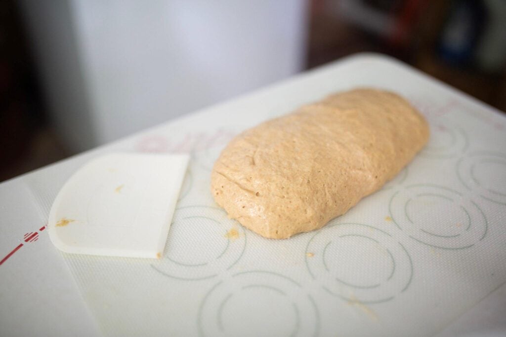 There is a piece of dough shaped into a log on a silicone mat beside a dough scraper.