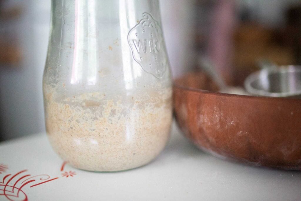 A jar of sourdough starter is beside a dark wood bowl with a measuring cup in it.