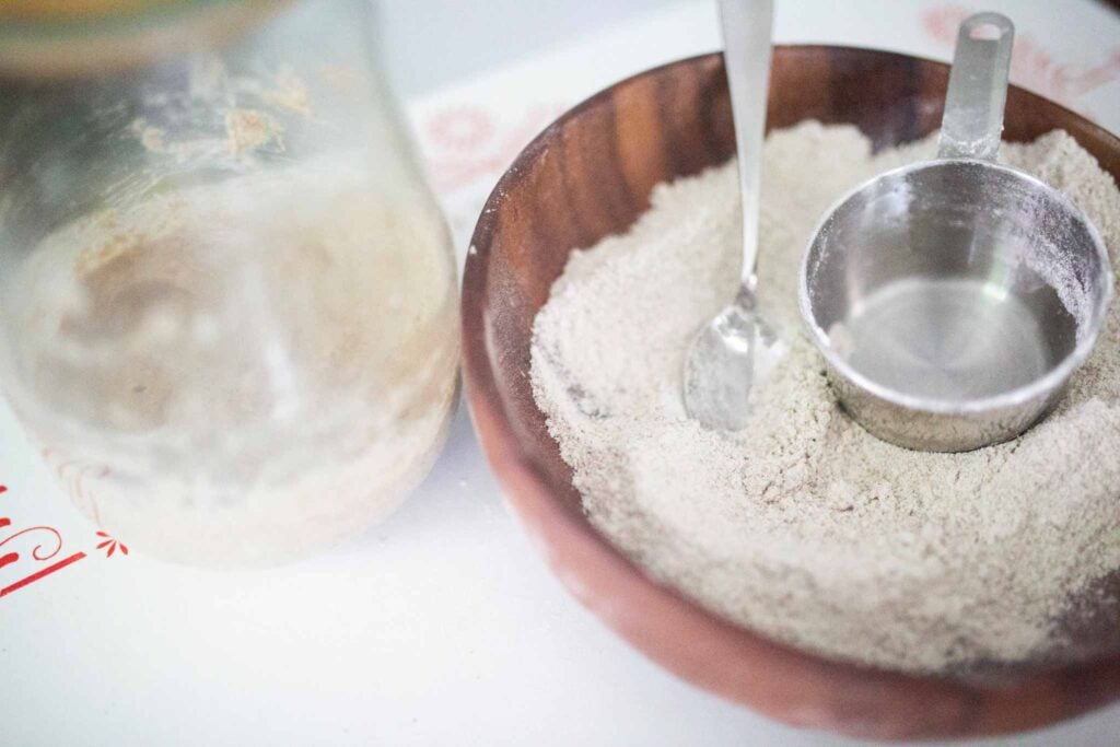 There is a jar with a small amount of sourdough starter in it beside a dark wood bowl full of flour with a spoon and measuring cup.