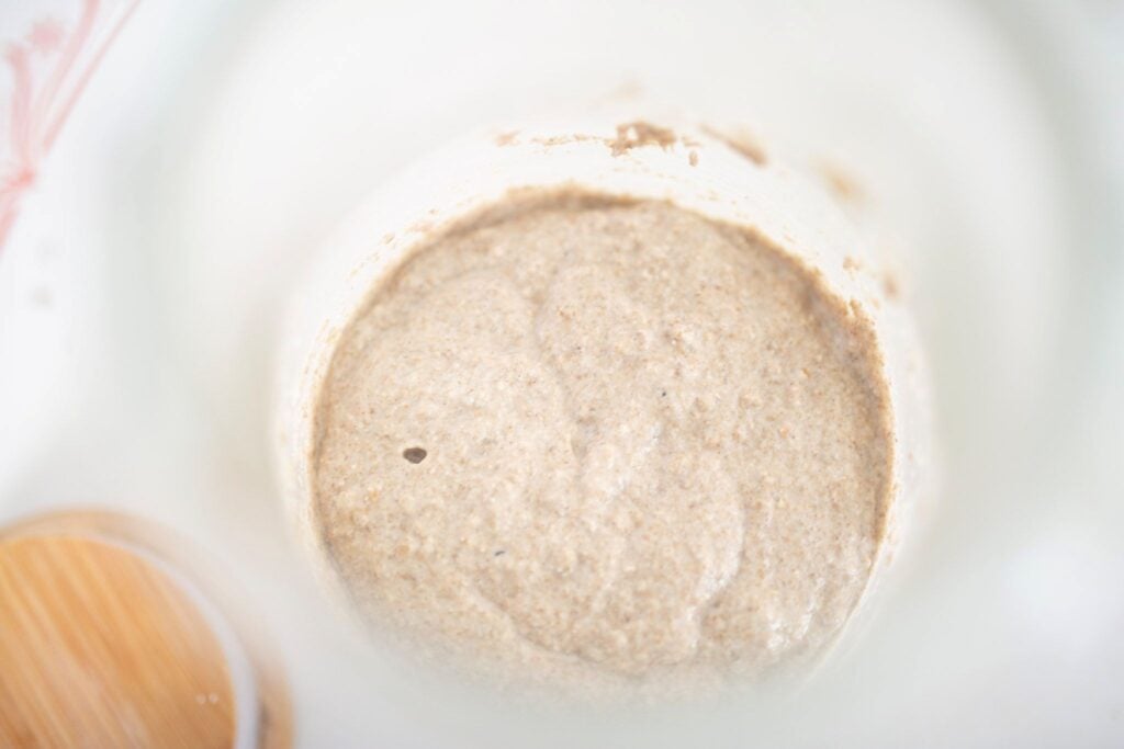 Overhead photo looking into a jar of sourdough starter. There is one bubble on the surface and wooden lid laid beside the jar.