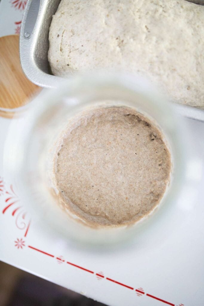 This is an overhead photo of sourdough starter in a jar beside a loaf pan of bread dough.