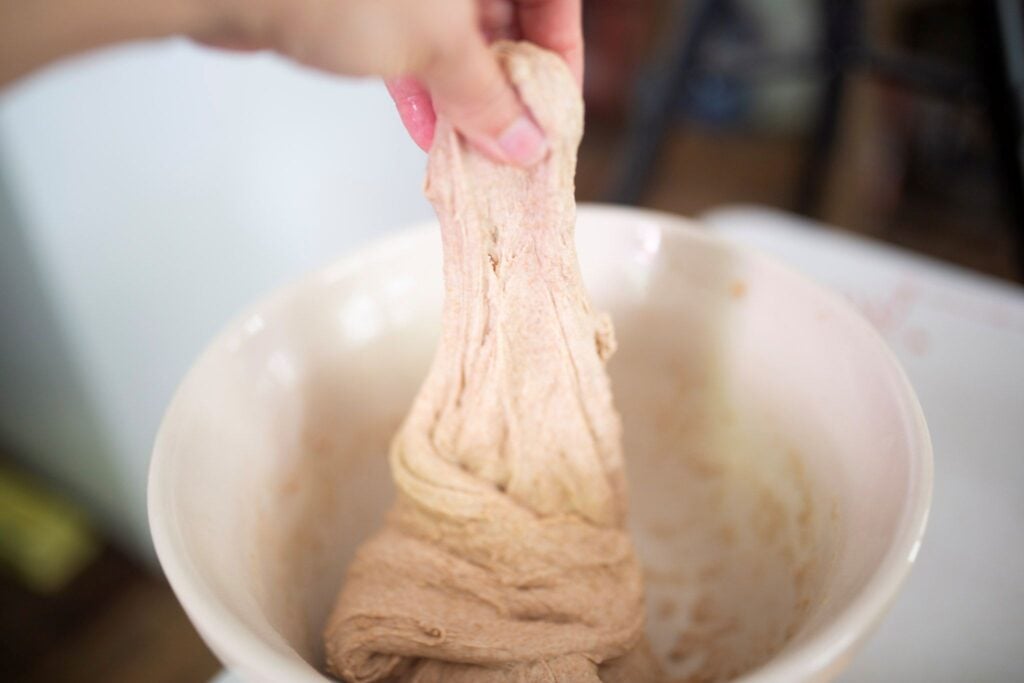 A hand stretching dough up in the air that is in a cream colored bowl.