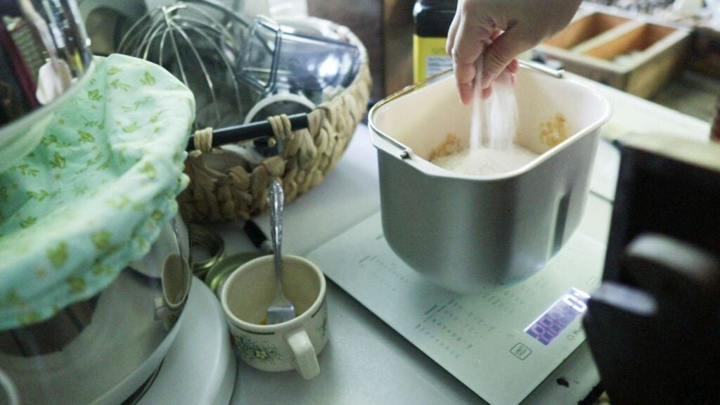 A hand is adding salt to a bread machine loaf pan.
