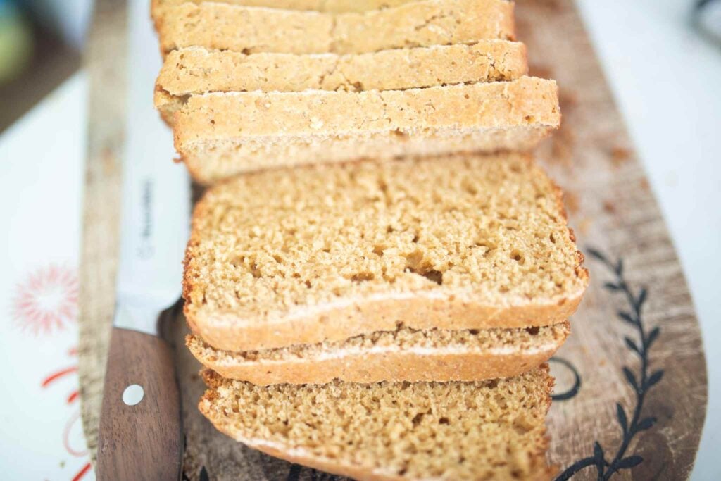 A loaf of sandwich bread sliced on a cutting board beside a bread knife with a wooden handle.