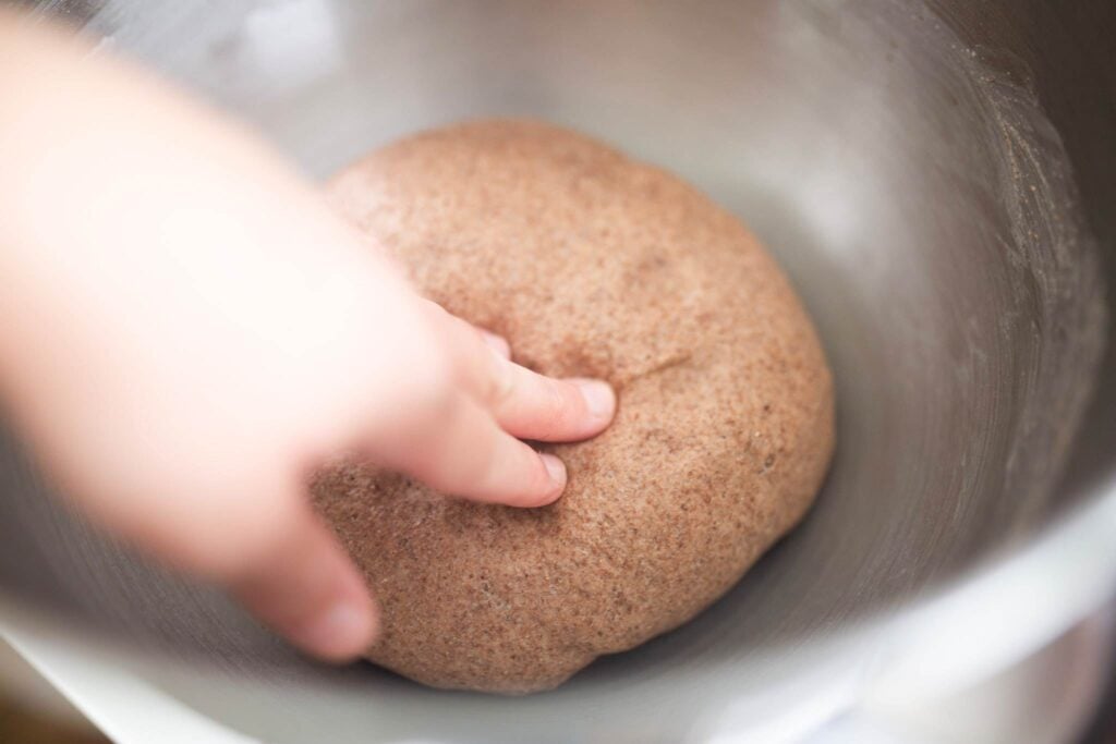 A hand is pressing on risen bread dough in a silver bowl.
