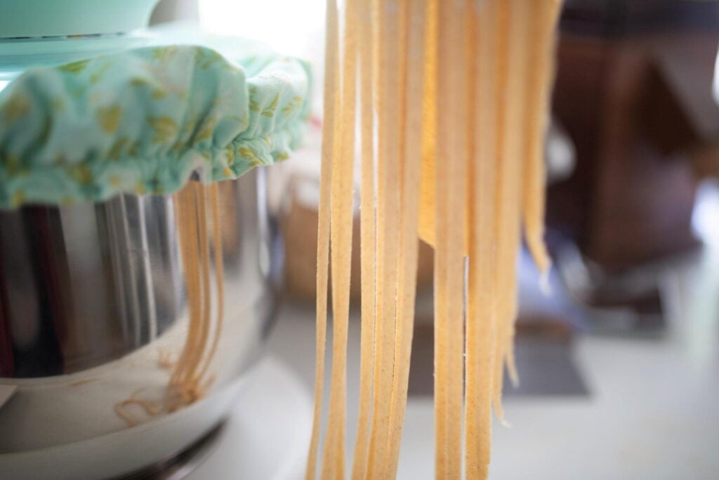 Pasta noodles hanging from pasta maker with mixer bowl in the background.