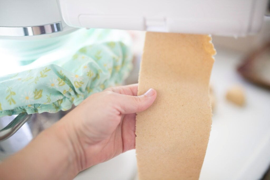 A hand is holding a pasta sheet hanging from a pasta maker.