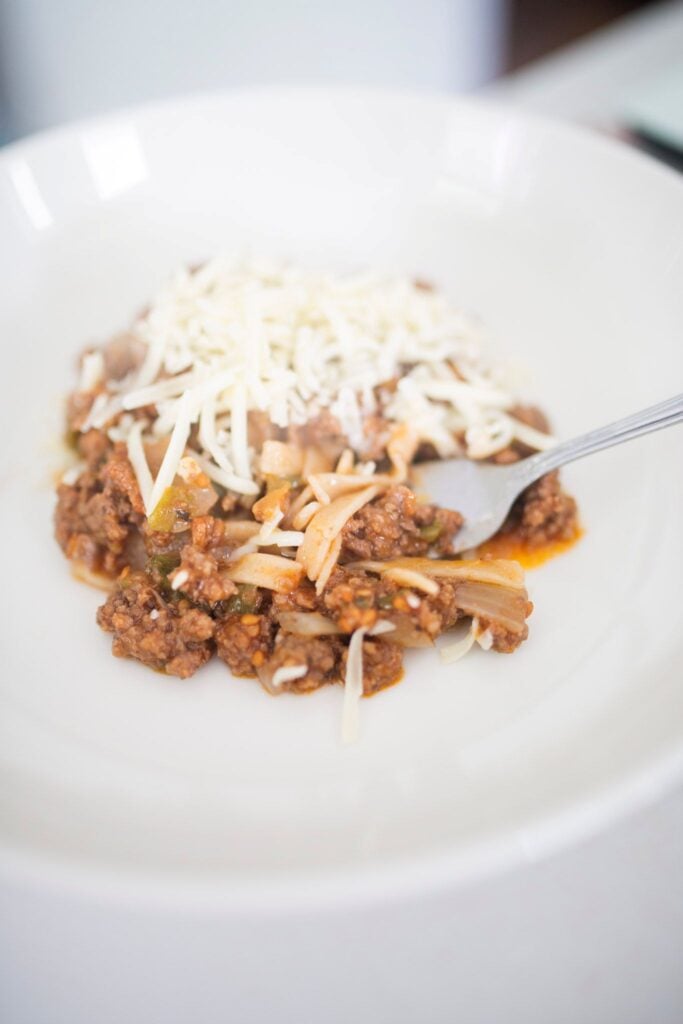 Pasta noodles are topped with a meat and tomato sauce and shredded cheese is on a white plate.