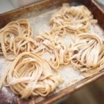 Mounds of fresh pasta noodles are laying on a floured baking sheet.