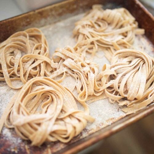 Mounds of fresh pasta noodles are laying on a floured baking sheet.
