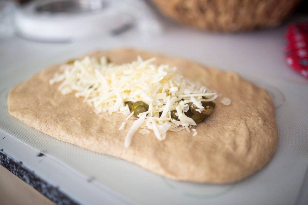 A rectangle of dough is topped with shredded cheese and peppers on a silicone dough mat.