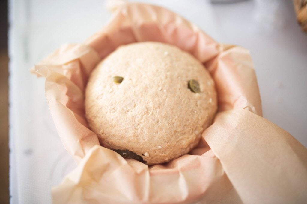 A ball of bread dough with jalapenos showing through sits in a bowl lined with parchment paper.