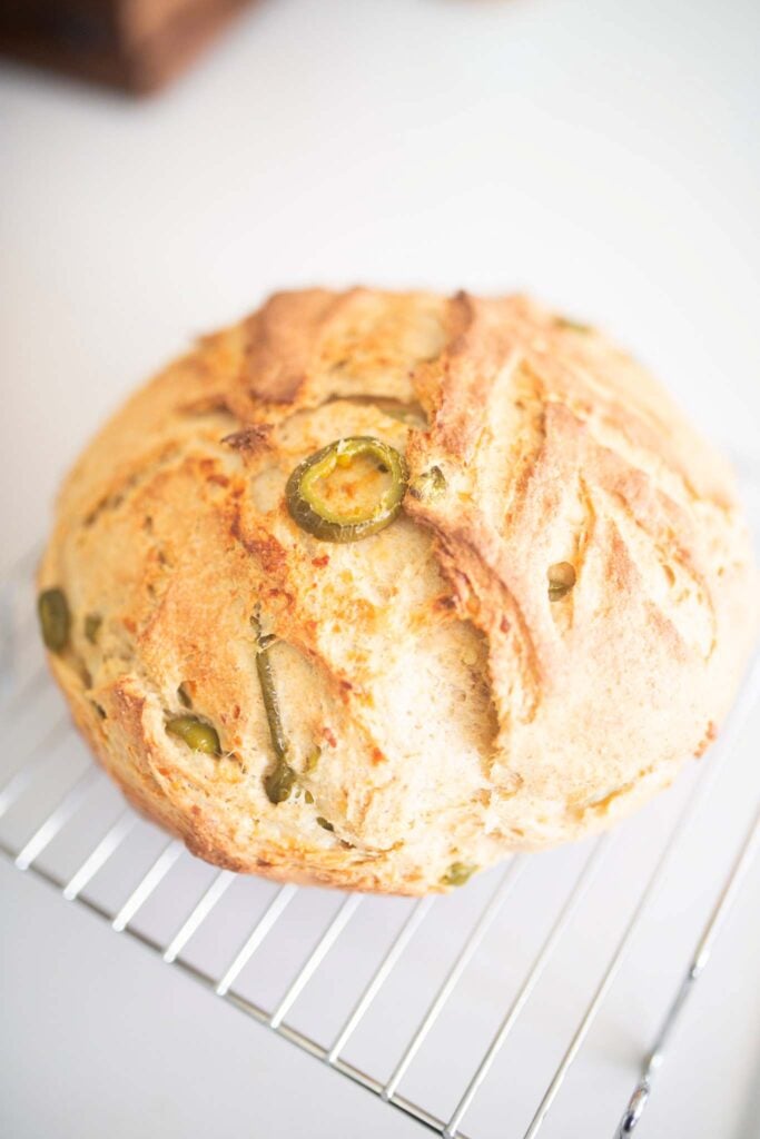 An artisan loaf with a jalapeno slice on top sits on a wire rack on a white table.