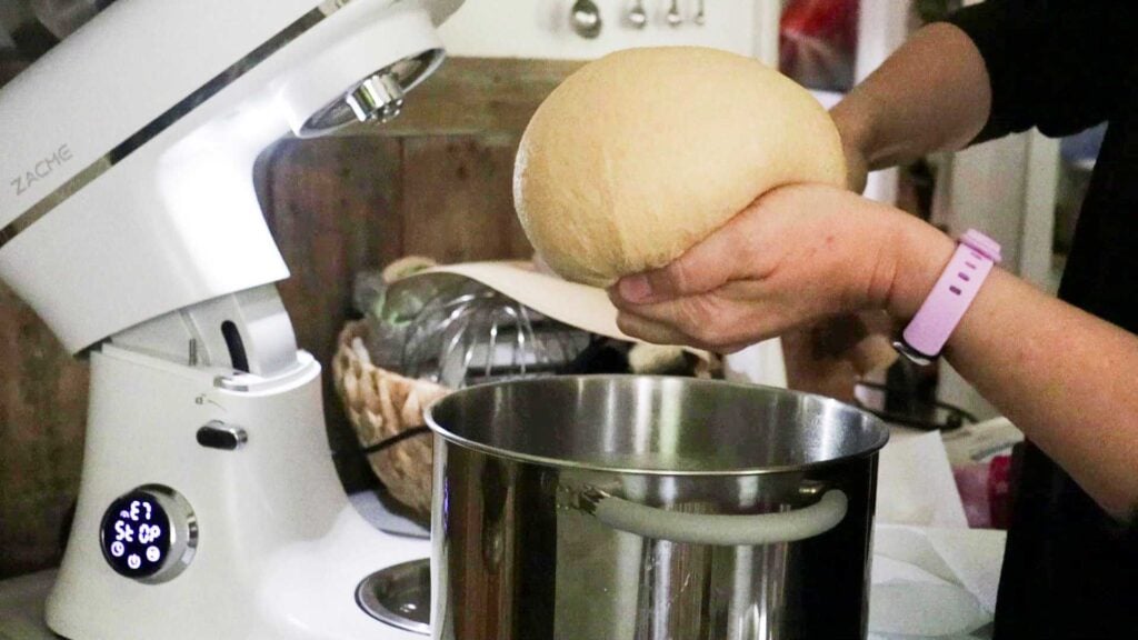 Hands are holding a ball of kneaded dough over a stainless steel bowl.